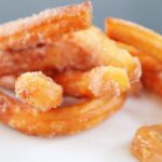 Plate of golden homemade churros coated in cinnamon sugar with a side of caramel dipping