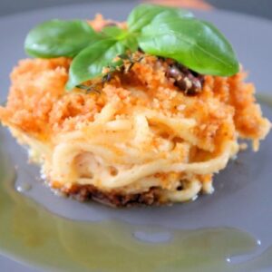 Golden baked leftover pasta with melted cheese, olives, and herbs served in a black plated drizzled with olive oil
